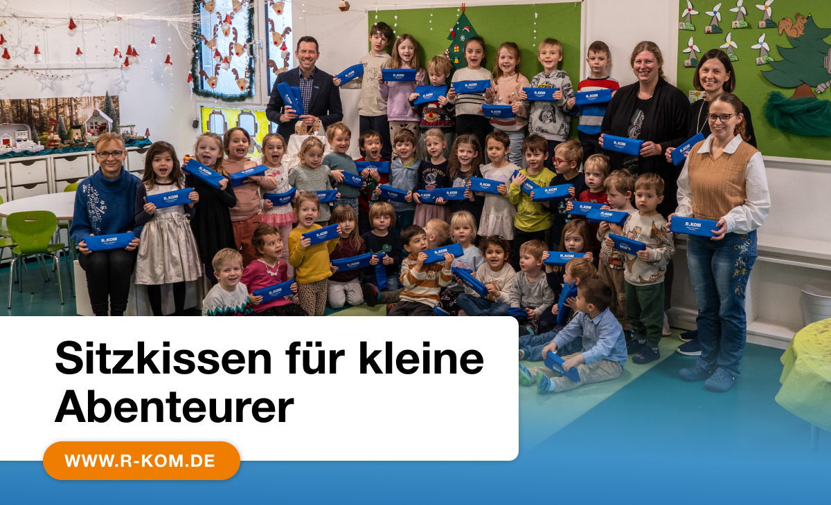 Gruppe von Kindern und Erziehern in einer Kita, die blaue R-KOM-Sitzkissen halten, mit dem Text ‚Sitzkissen für kleine Abenteurer‘ und www.r-kom.de unten.