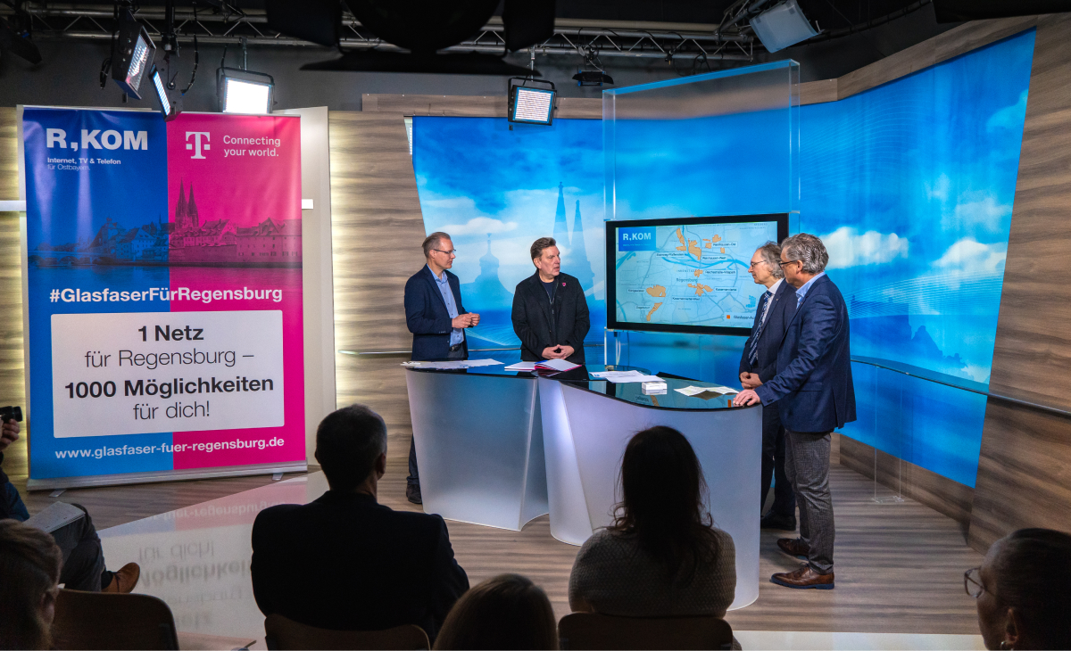 Pressekonferenz im Studio: Vertreter von R-KOM und der Telekom präsentieren Glasfaser-Initiative für Regensburg. Im Hintergrund eine Bildschirmpräsentation mit Netzplänen und ein Roll-Up mit der Aufschrift ‚#GlasfaserFürRegensburg: 1 Netz für Regensburg – 1000 Möglichkeiten für dich!‘. Publikum sitzt im Vordergrund.